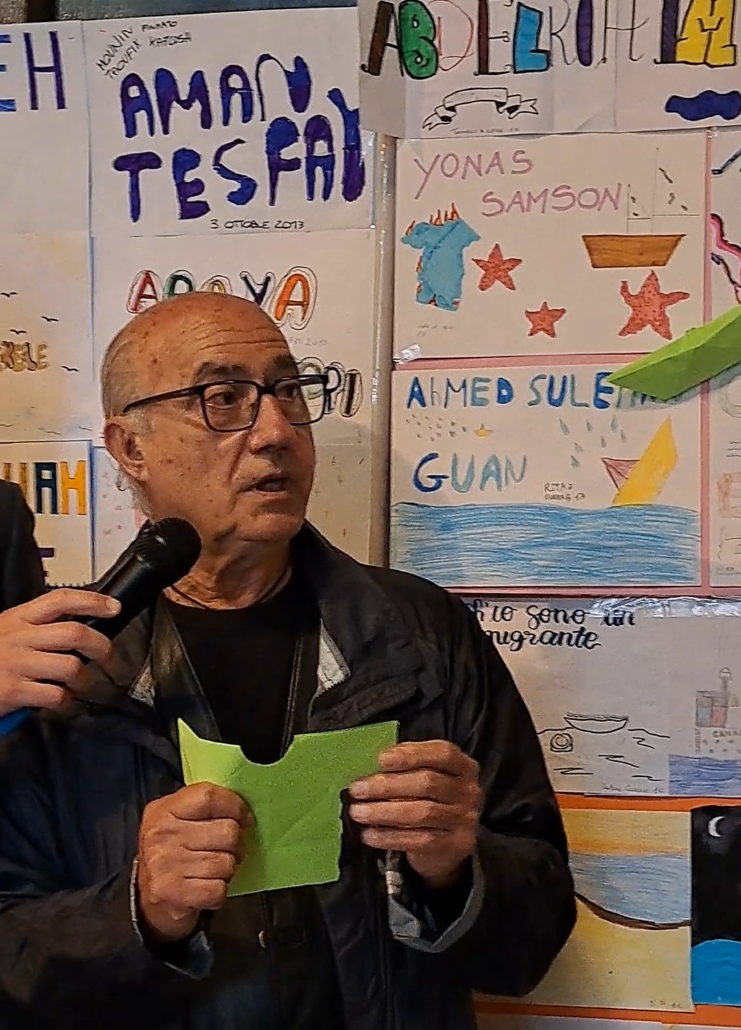 Vito Fiorino mostra il simbolo di carta ricavato tagliando una barchetta, alle sue spalle i disegni dei ragazzi con i nomi delle vittime del 3 ottobre 2013